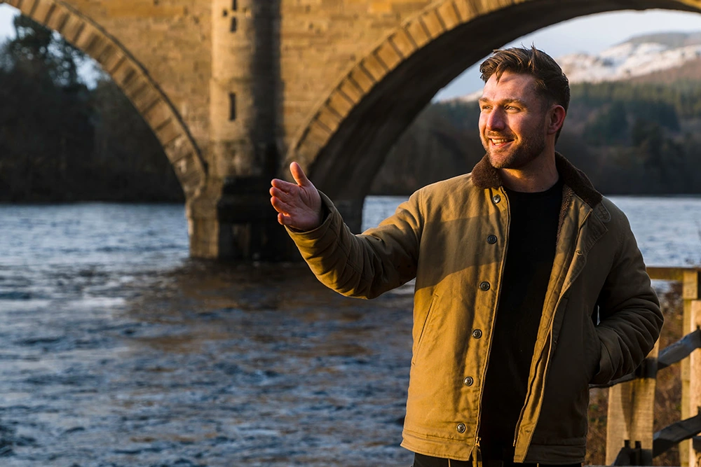 Ally Heather in foreground by the side of a river with a bridge in the background