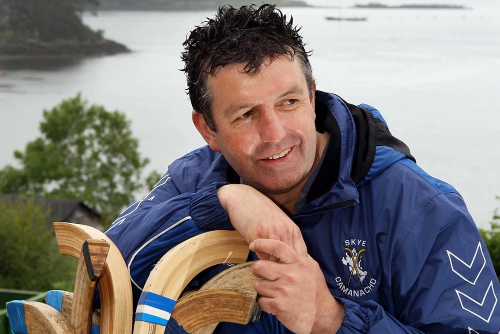 Lucky2BHere founder, the late Ross Cowie wearing a blue Skye Camanachd jacket leaning on a bag full of shinty sticks with Portree Bay, Isle of Skye in the background