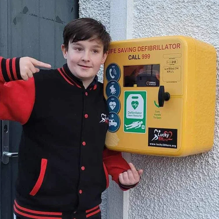 Young boy points to Lucky2BHere Defibrillato strategically placed on an a rural building exterior wall on Isle of Skye